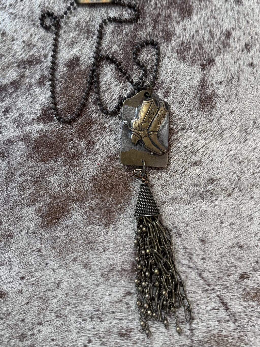 A brass antiqued finish metal necklace with a pendant featuring a cowboy hat design, attached to a chain of metal fringe.