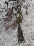 A brass antiqued finish metal necklace with a pendant featuring a cowboy hat design, attached to a chain of metal fringe.
