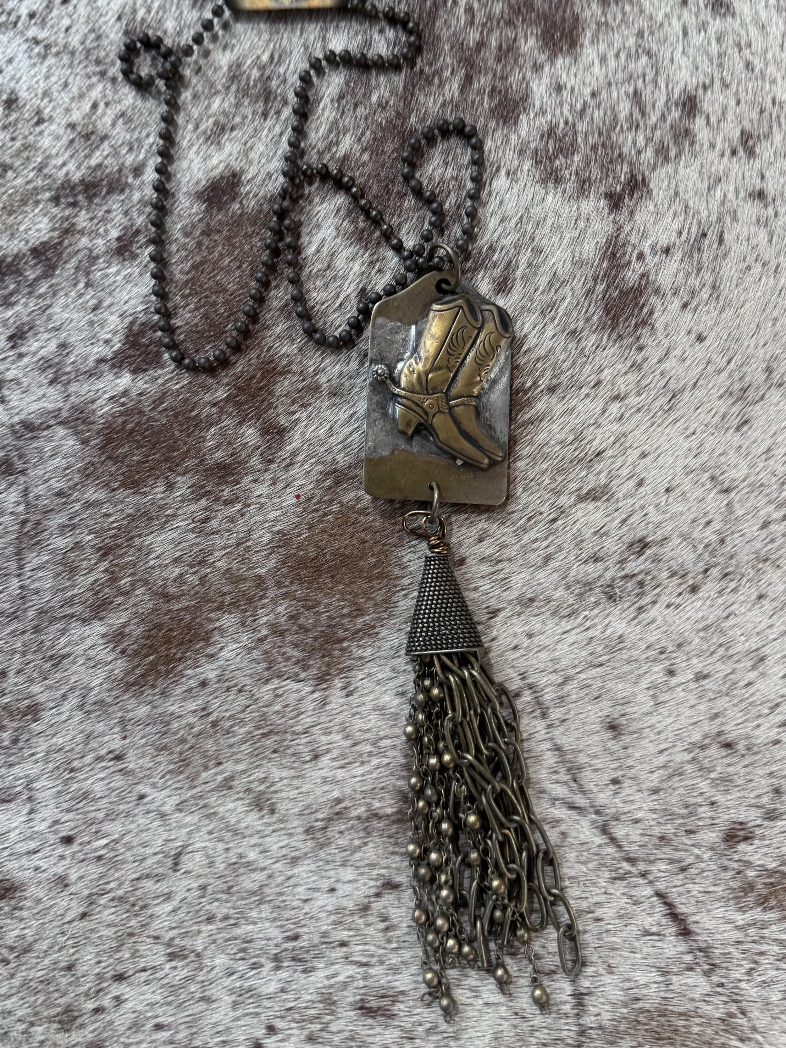 A brass antiqued finish metal necklace with a pendant featuring a cowboy hat design, attached to a chain of metal fringe.